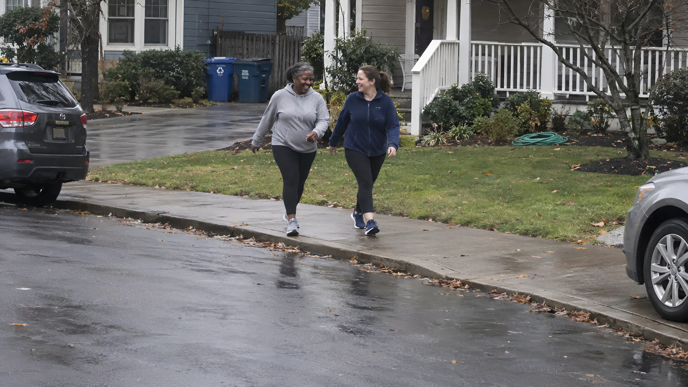 Two women walking fast and laughing together on a neighborhood sidewalk on a gray morning